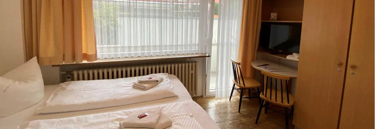 Blick von einem Appartment Richtung Fenster und Balkon. Auf dem Bett liegen die Handtücher zusammengefaltet auf der Bettdecke, neben dem Schrank befindet siche ine kleiner Tisch mit den wichtigsten Gästeinformationen. Über dem Tisch auf einem Regalboden steht der Flachbildschirm TV mit Smart Funktion.