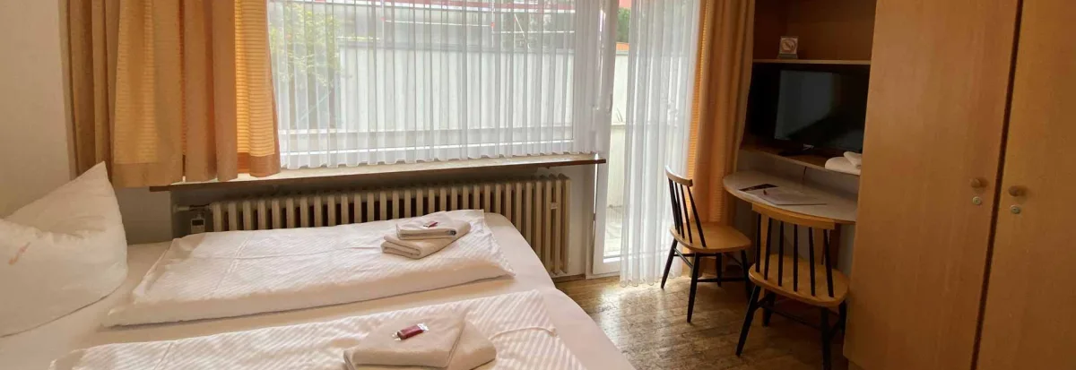 Blick von einem Appartment Richtung Fenster und Balkon. Auf dem Bett liegen die Handtücher zusammengefaltet auf der Bettdecke, neben dem Schrank befindet siche ine kleiner Tisch mit den wichtigsten Gästeinformationen. Über dem Tisch auf einem Regalboden steht der Flachbildschirm TV mit Smart Funktion.